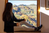 A woman in casual attire enjoys coffee while sitting by a window with a scenic view.