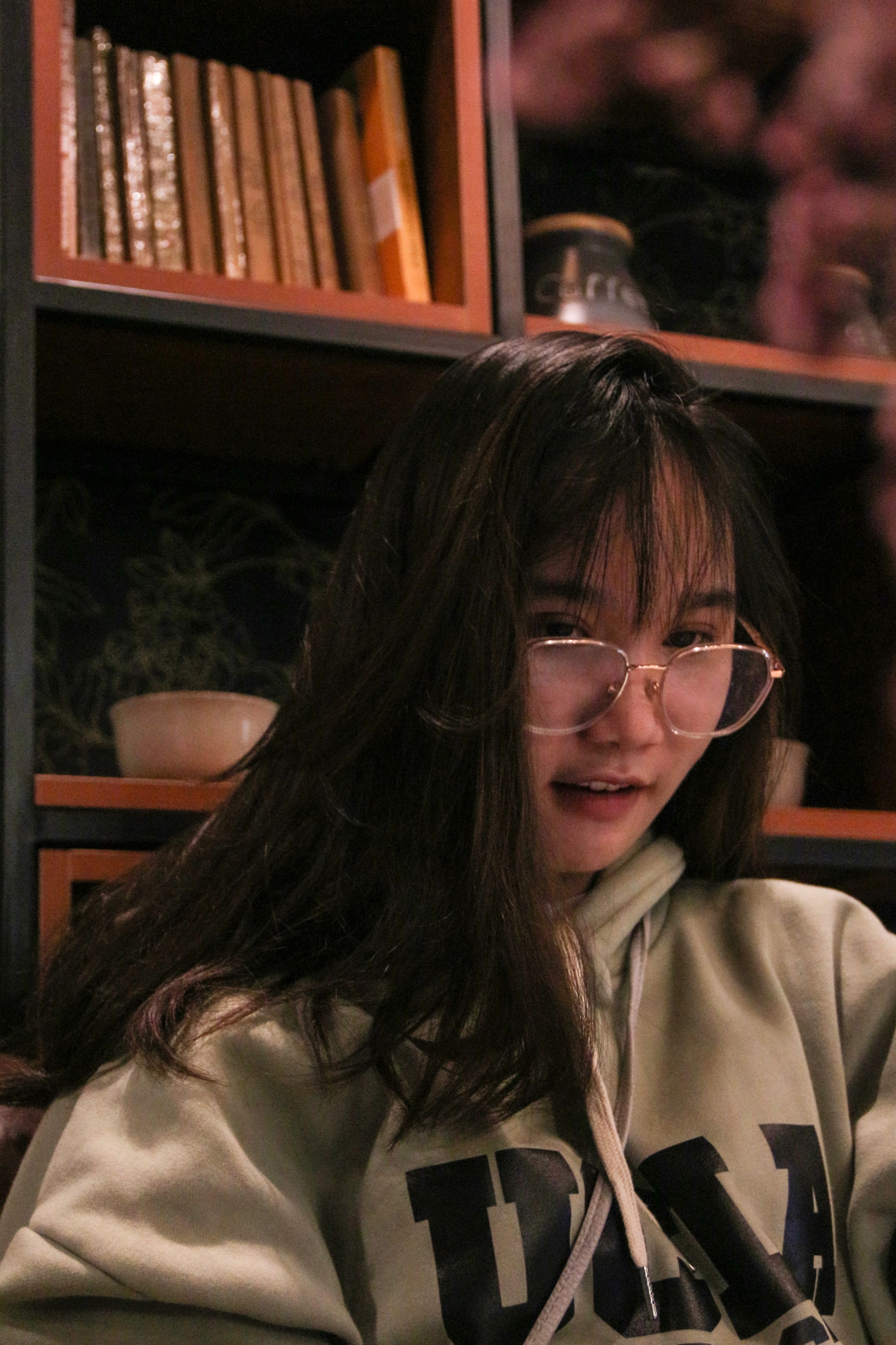 Asian woman in glasses, wearing a hoodie, relaxing indoors with a bookshelf behind her, creating a cozy ambiance.