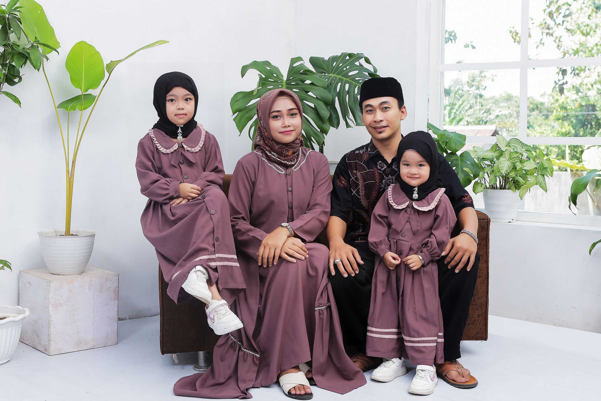 A family of four sitting together in matching hijabs and dresses, indoors with potted plants.