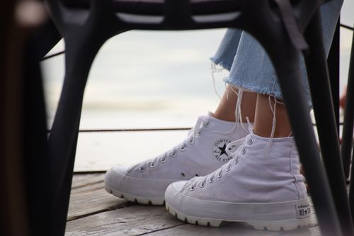 Free White sneakers paired with denim jeans on a wooden deck, viewed through a chair. Stock Photo