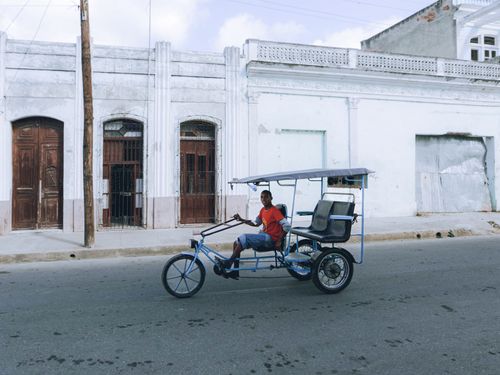 Free An adult man riding a colorful tricycle taxi on a quiet urban street with historic buildings. Stock Photo
