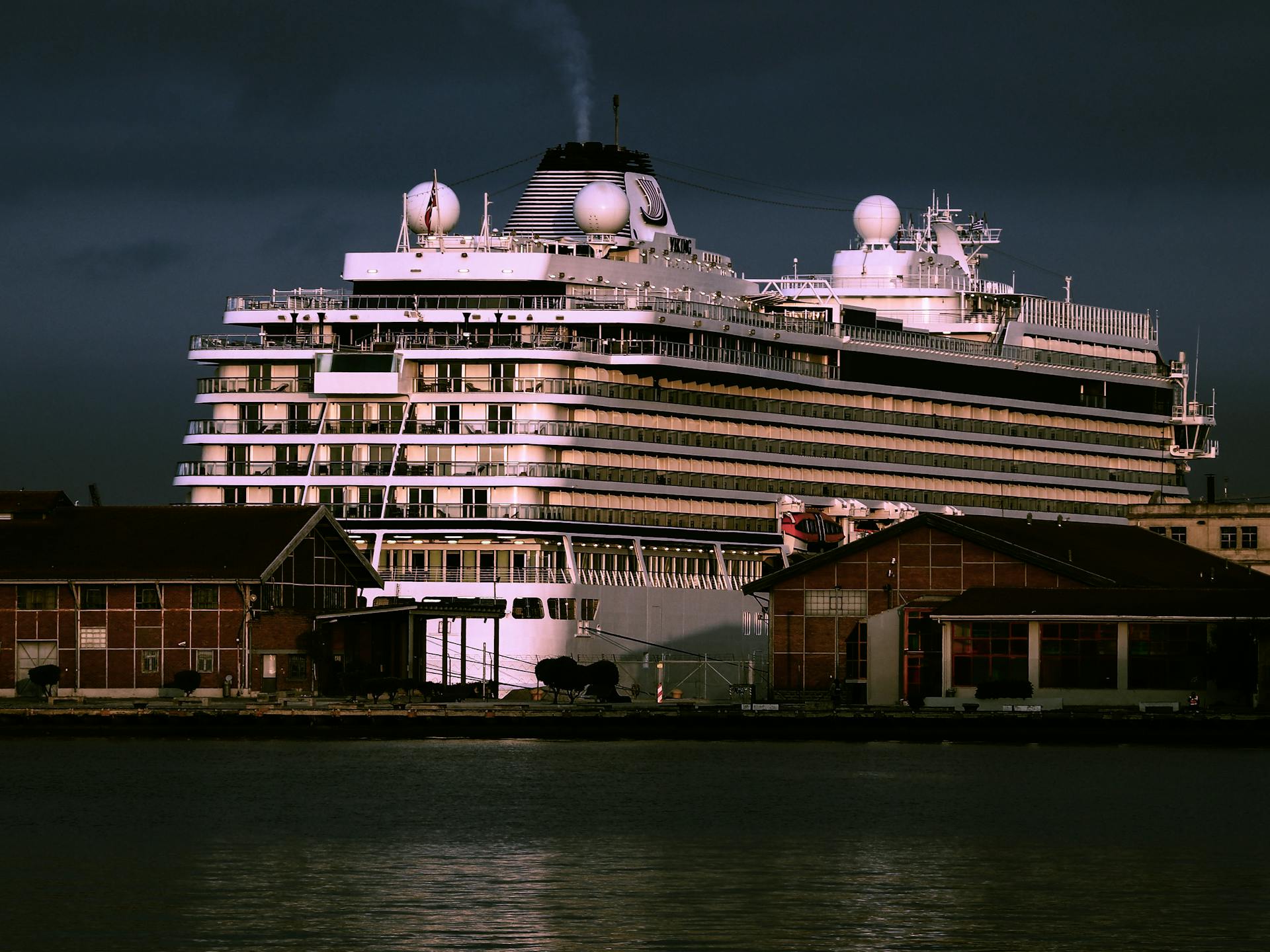 A large luxury cruise ship docked at a harbor, illuminated against the evening sky.