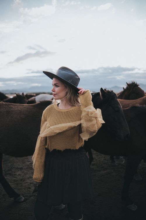 Free A woman wearing a hat stands among a group of horses in a natural setting, presenting a blend of fashion and nature. Stock Photo