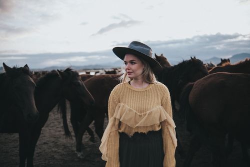 Free Woman in a stylish outfit and hat standing among a herd of horses outdoors at dusk. Stock Photo
