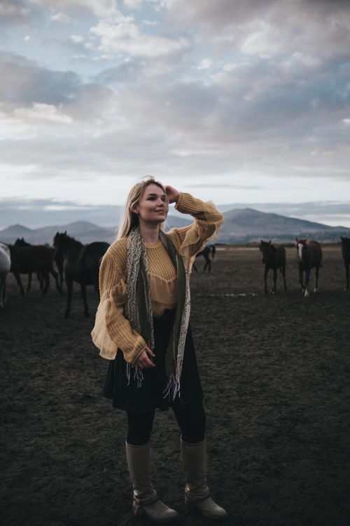 Free A woman in a beige sweater stands in a pastoral landscape with horses under an overcast sky. Stock Photo