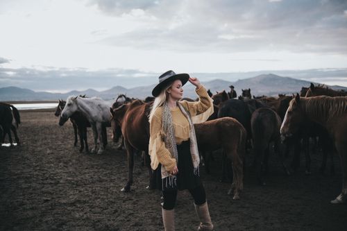 Free Blonde woman in a hat posing among horses in a picturesque landscape. Stock Photo