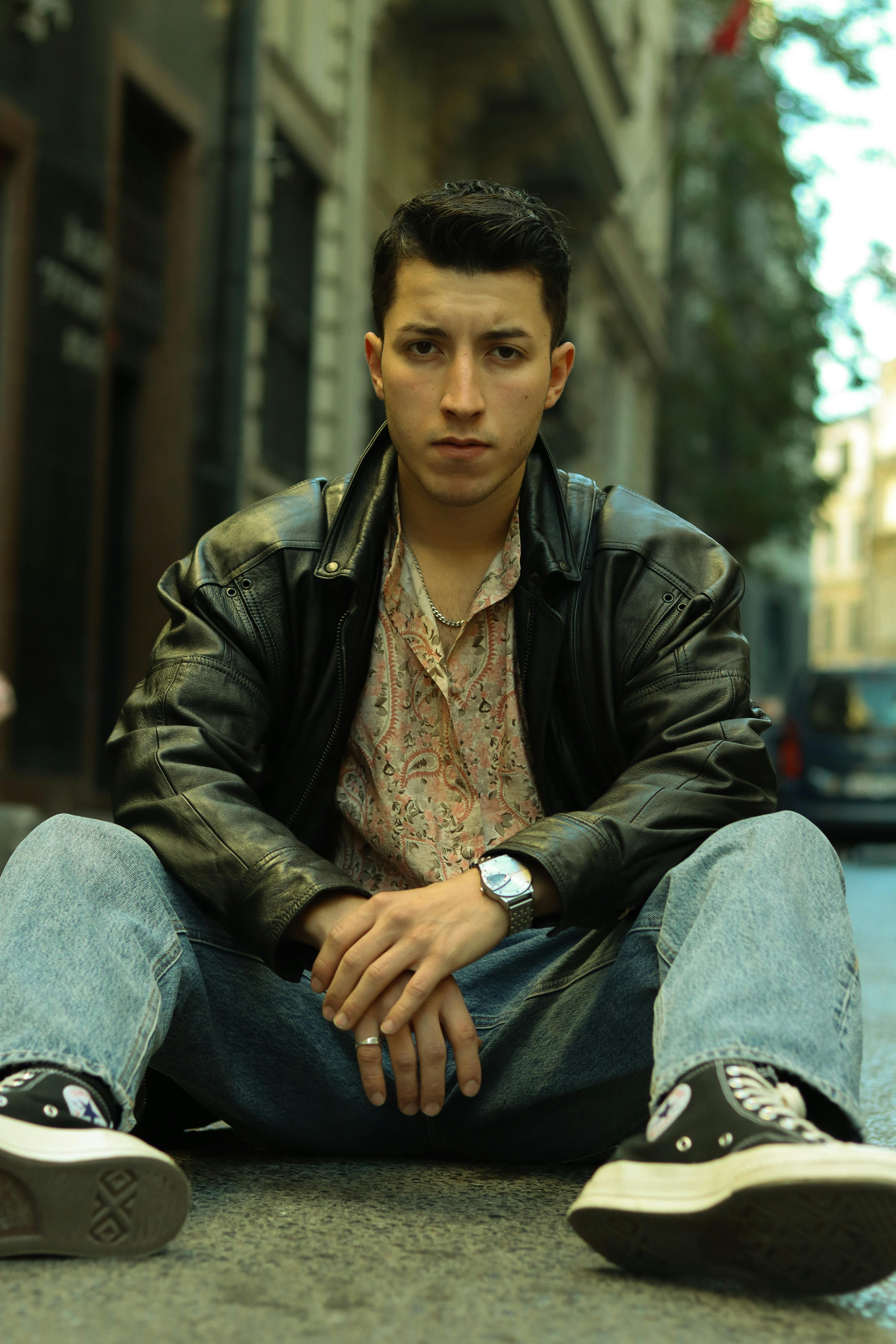 Man sitting in an urban setting wearing a leather jacket and jeans.
