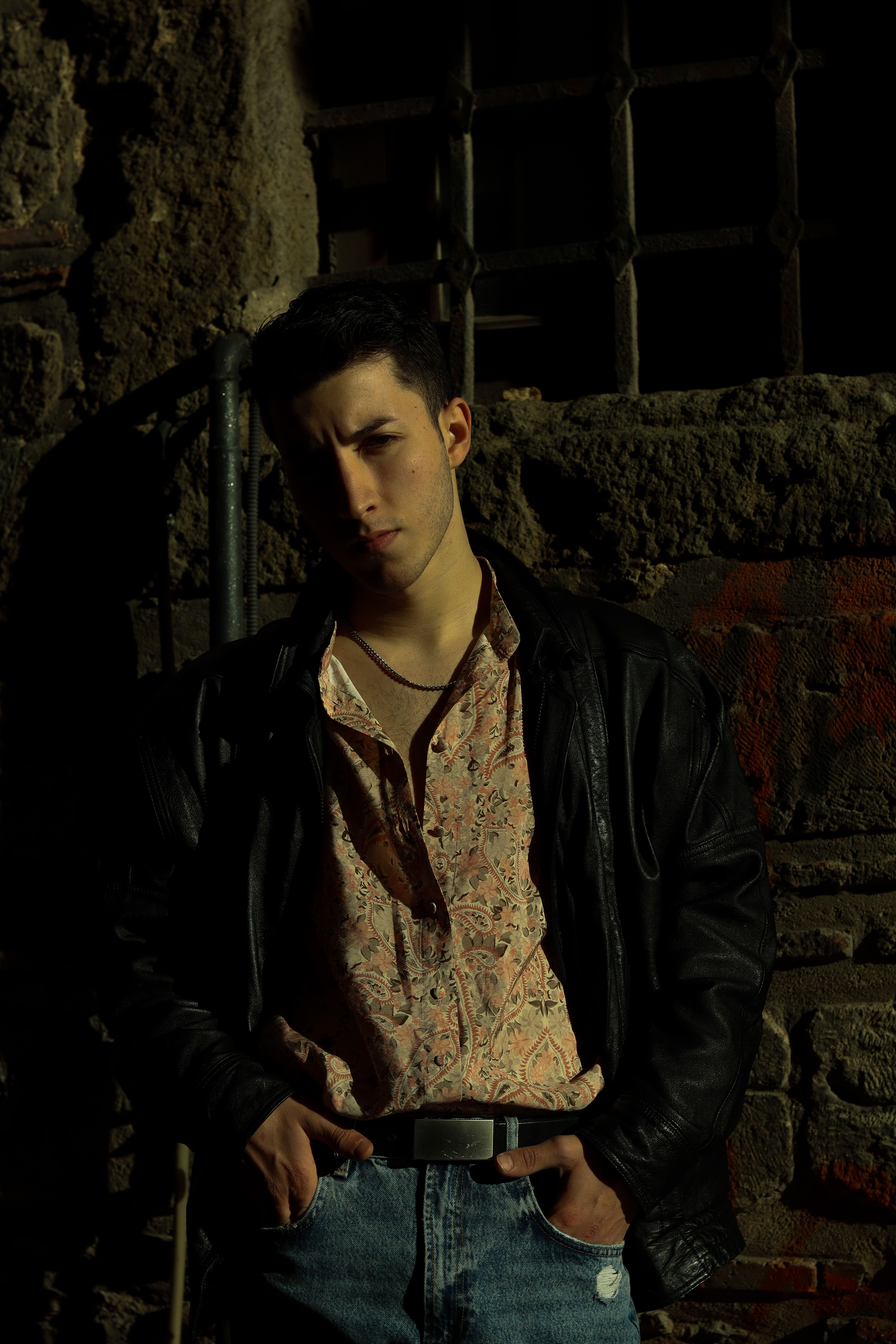 A man stands in a shadowy environment, exuding style with a black leather jacket and jeans.