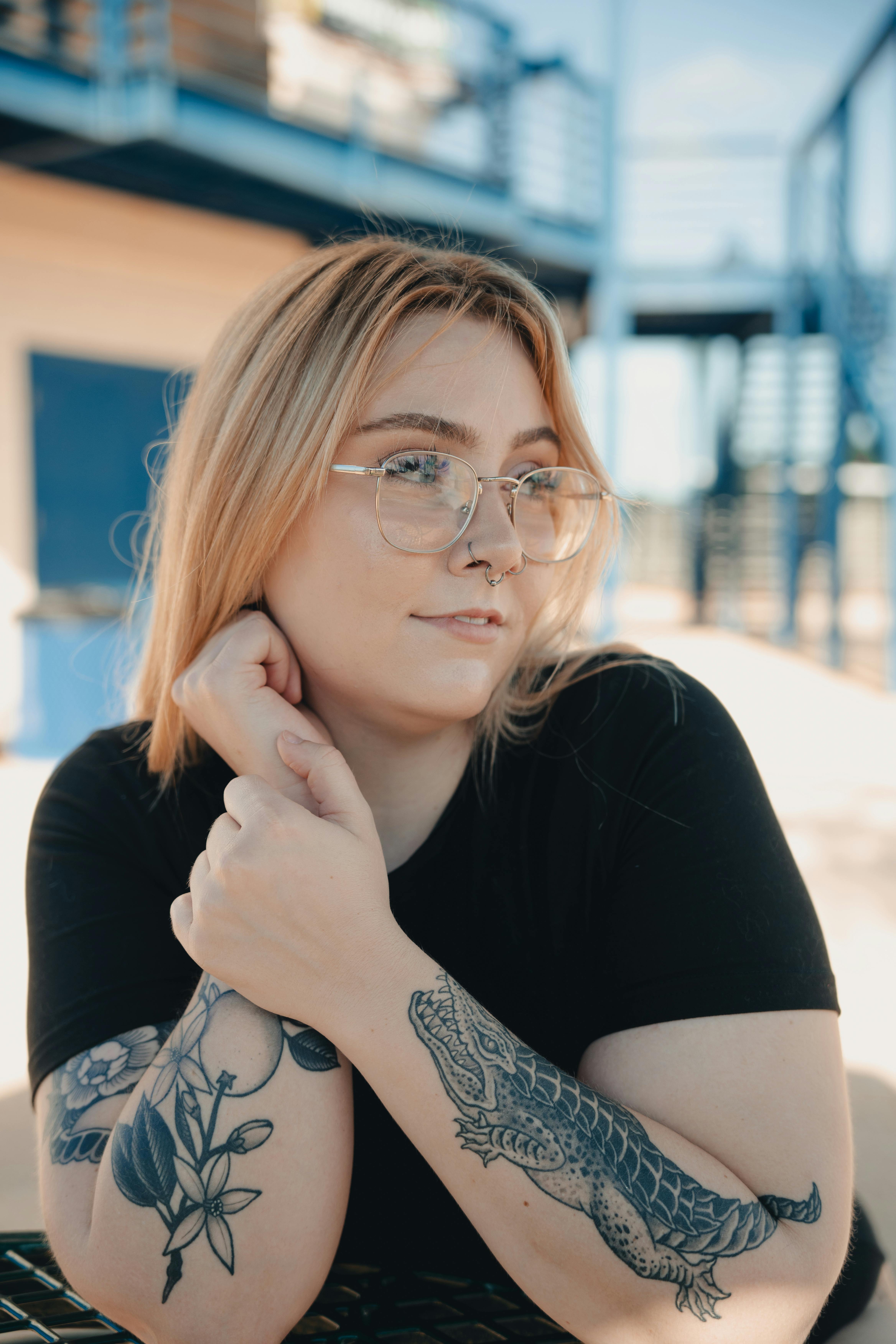 Caucasian woman with arm tattoos and eyeglasses, posing outdoors during the day.