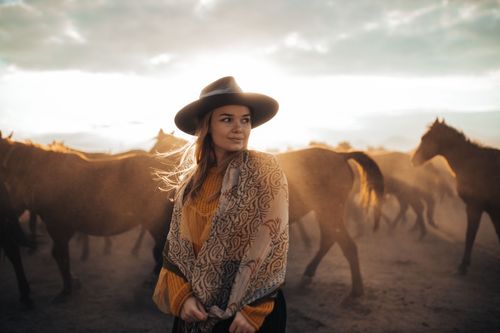 Free A woman in a cowboy hat and scarf stands in a field with horses in the background during sunset. Stock Photo