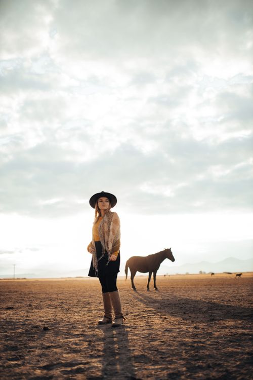 Free A woman in a cowboy hat stands in a vast open field under a cloudy sky with a horse nearby, capturing rustic lifestyle. Stock Photo