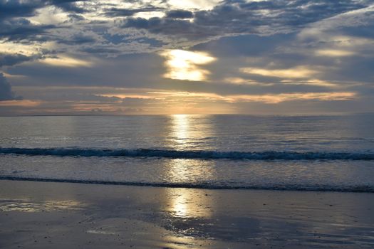 Peaceful beach scene at sunrise with gentle waves and a cloudy sky reflecting sunlight.
