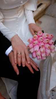 Bride and groom holding hands with a bouquet of pink tulips, showcasing wedding rings and elegant attire.