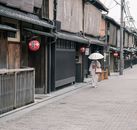 在日本京都的一条传统街道上，一名身穿和服的妇女正撑着雨伞行走
