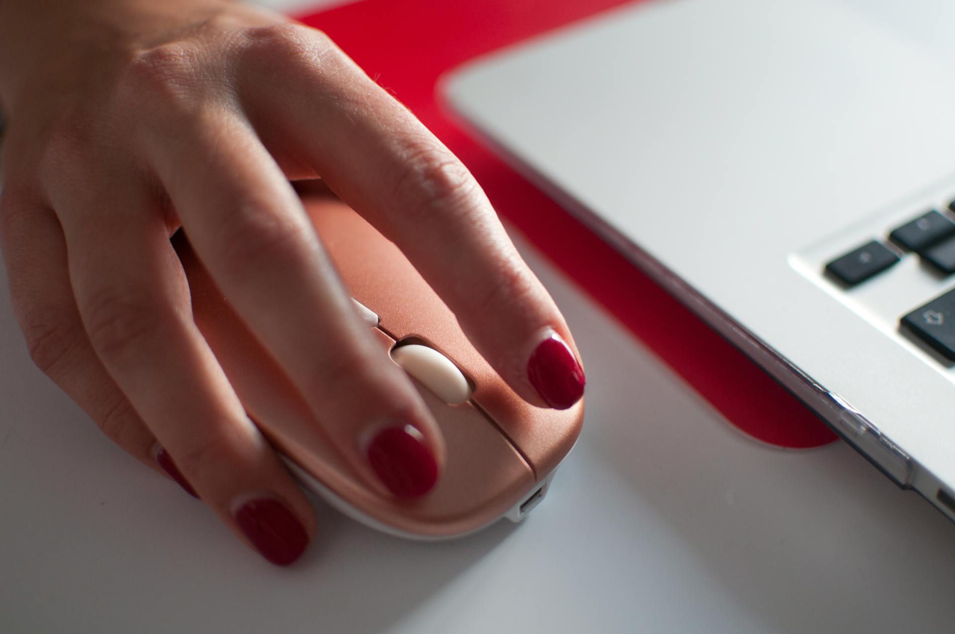 A woman's hand with painted nails using a stylish computer mouse beside a laptop.