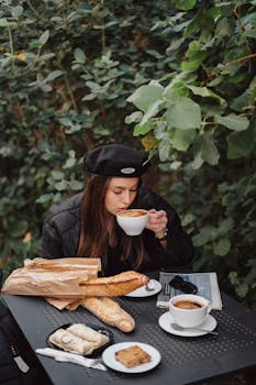 A woman sipping coffee in a French cafe setting with pastries and baguettes on a table.