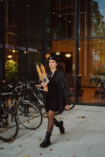A stylish woman holding baguettes walks past bicycles in an urban setting, exuding confidence and elegance.