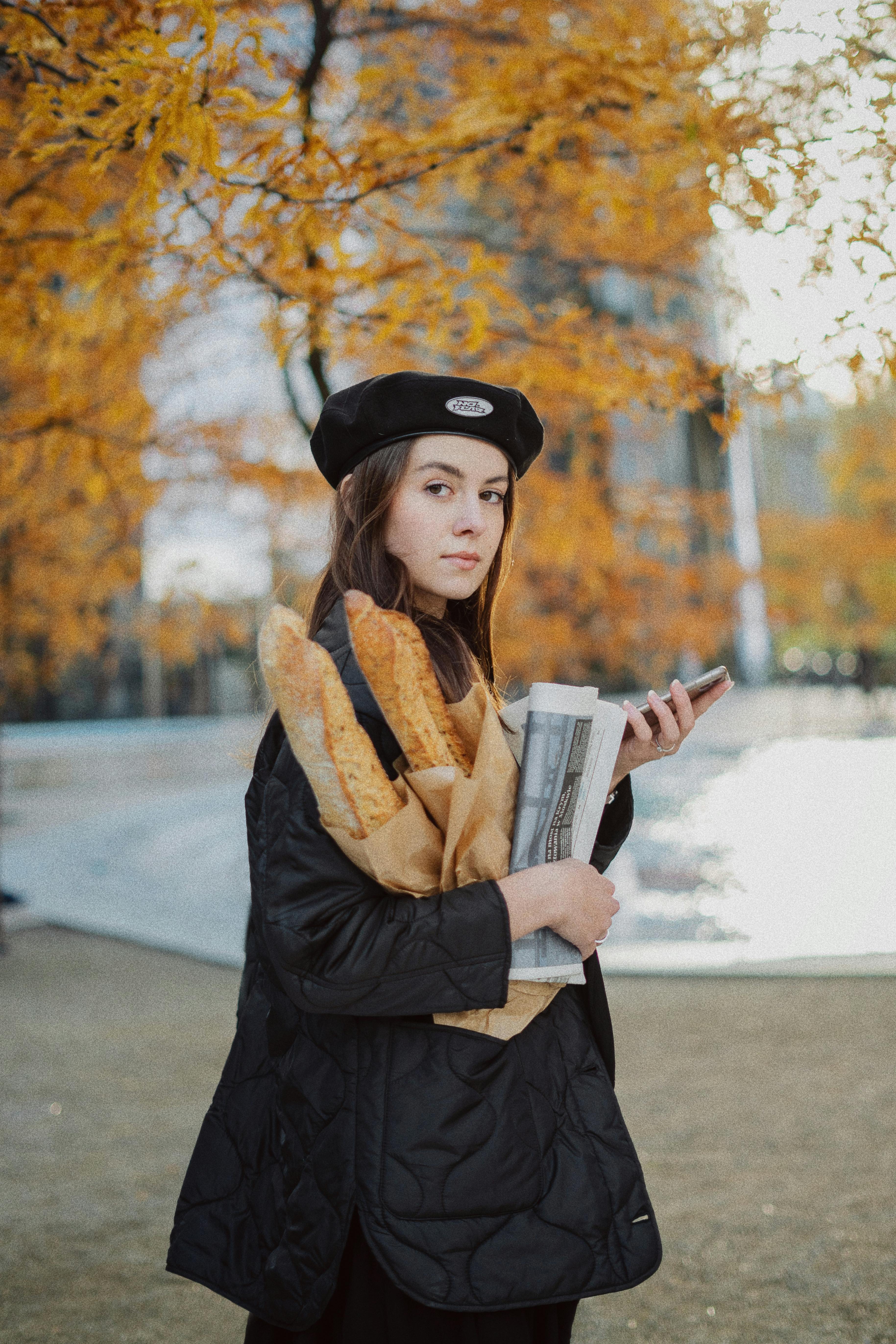 Stylish woman in a beret holding baguettes and newspaper during fall in a park, embodying French elegance.