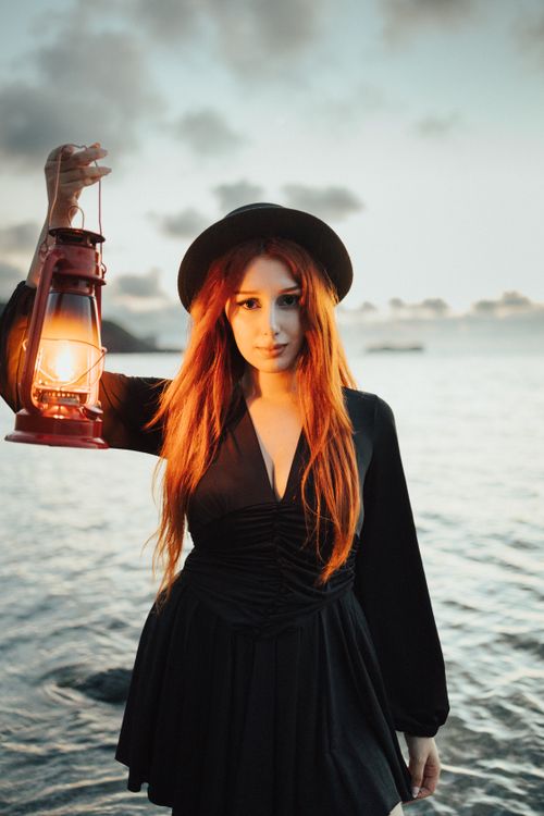 Free A woman in black dress and hat holds a lantern by the sea under a cloudy twilight sky. Stock Photo