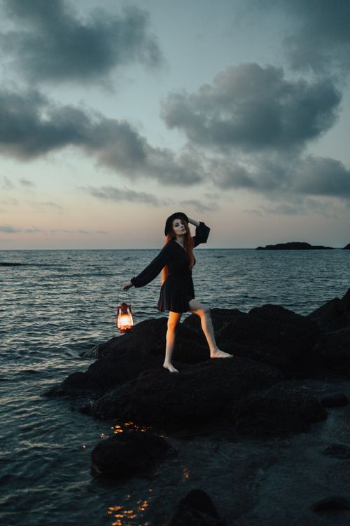 Free A woman holding a lantern stands on rocky shore at sunset, creating a moody and dramatic scene. Stock Photo