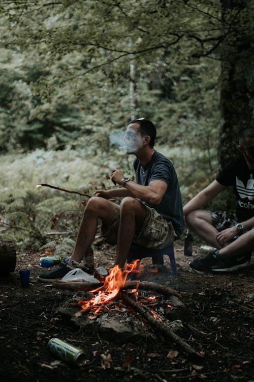 Man Smoking and Sitting on Stool Near Burning Wood