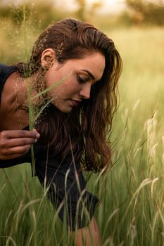 A woman with long brunette hair gently holds a plant stem in a grassy field.