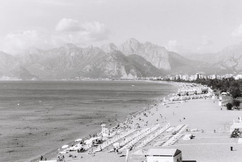 Grayscale Photo of People on the Sea Shore near Mountains