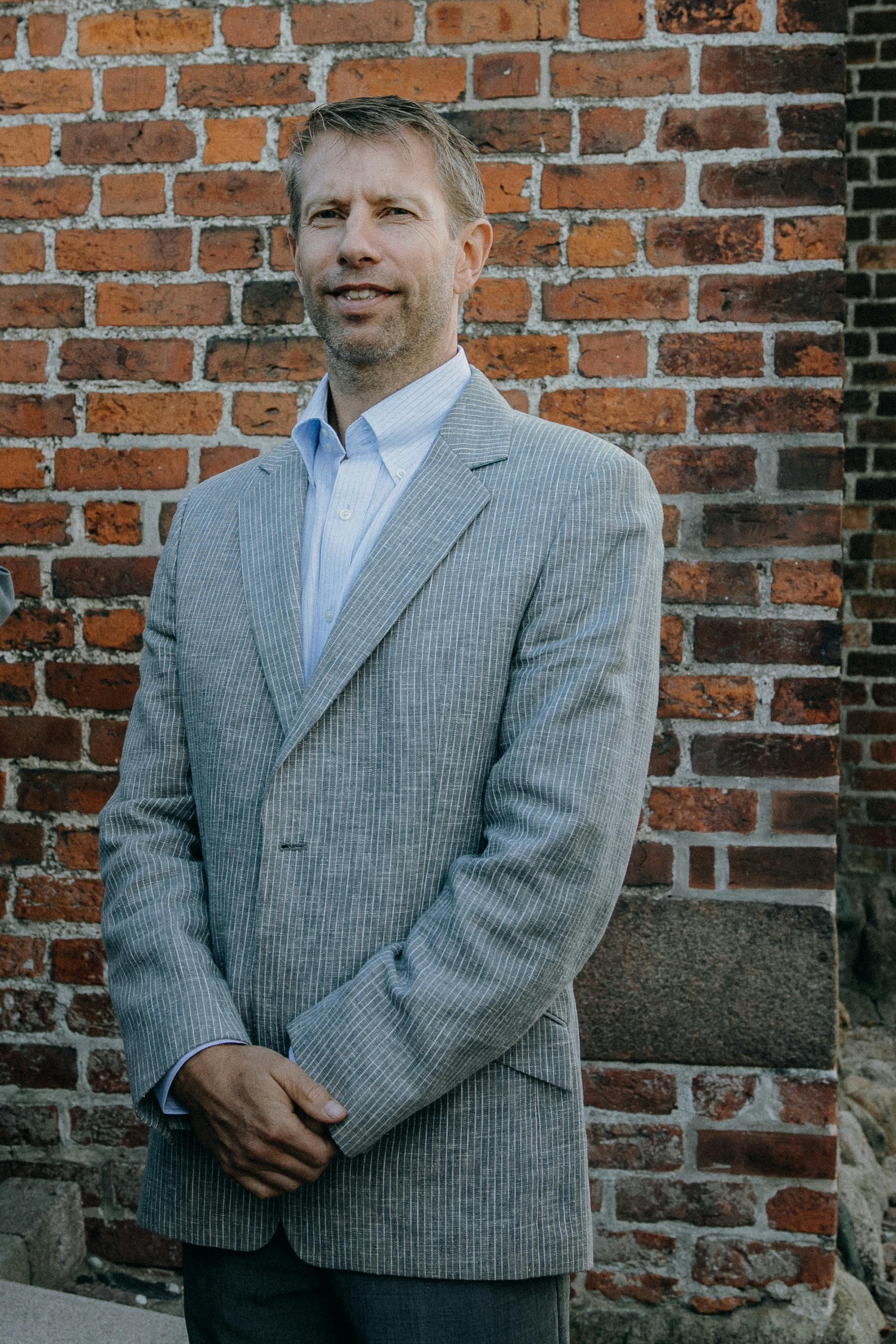 A man in a gray blazer stands confidently against a brick wall outdoors.