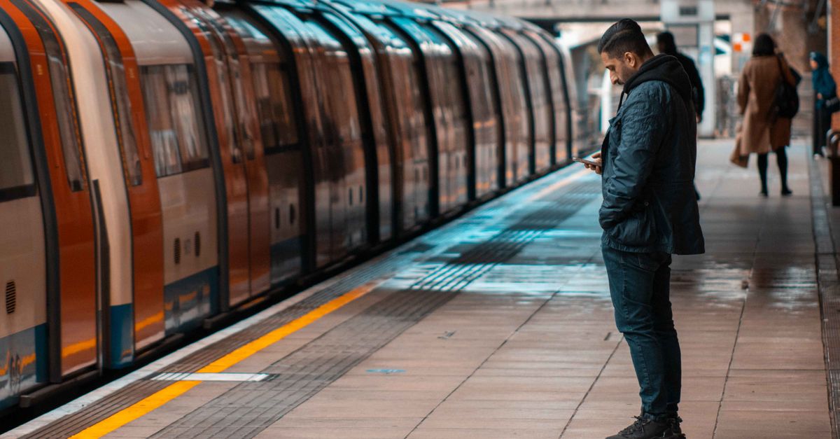 A man waits at a railway platform, texting on his cellphone, as a train arrives during daylight.
