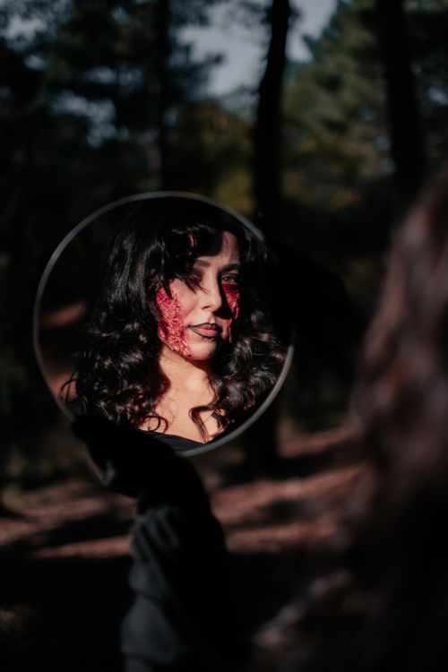 Free Spooky Halloween-themed portrait of a woman with creative bloody makeup looking into a mirror outdoors. Stock Photo