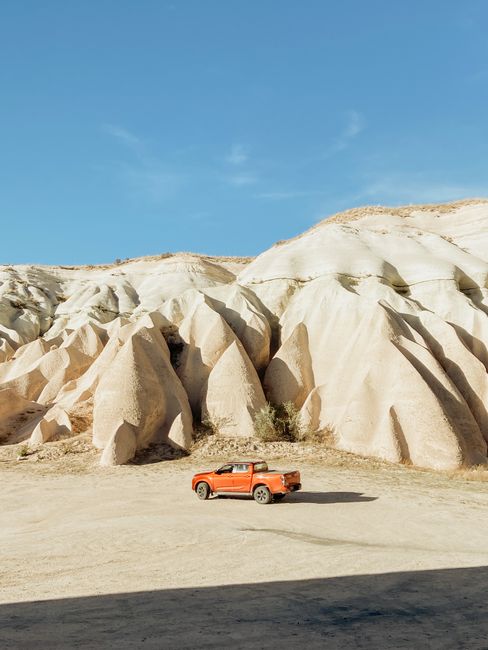 An orange SUV driving through an impressive desert landscape with unique rock formations under a clear blue sky.