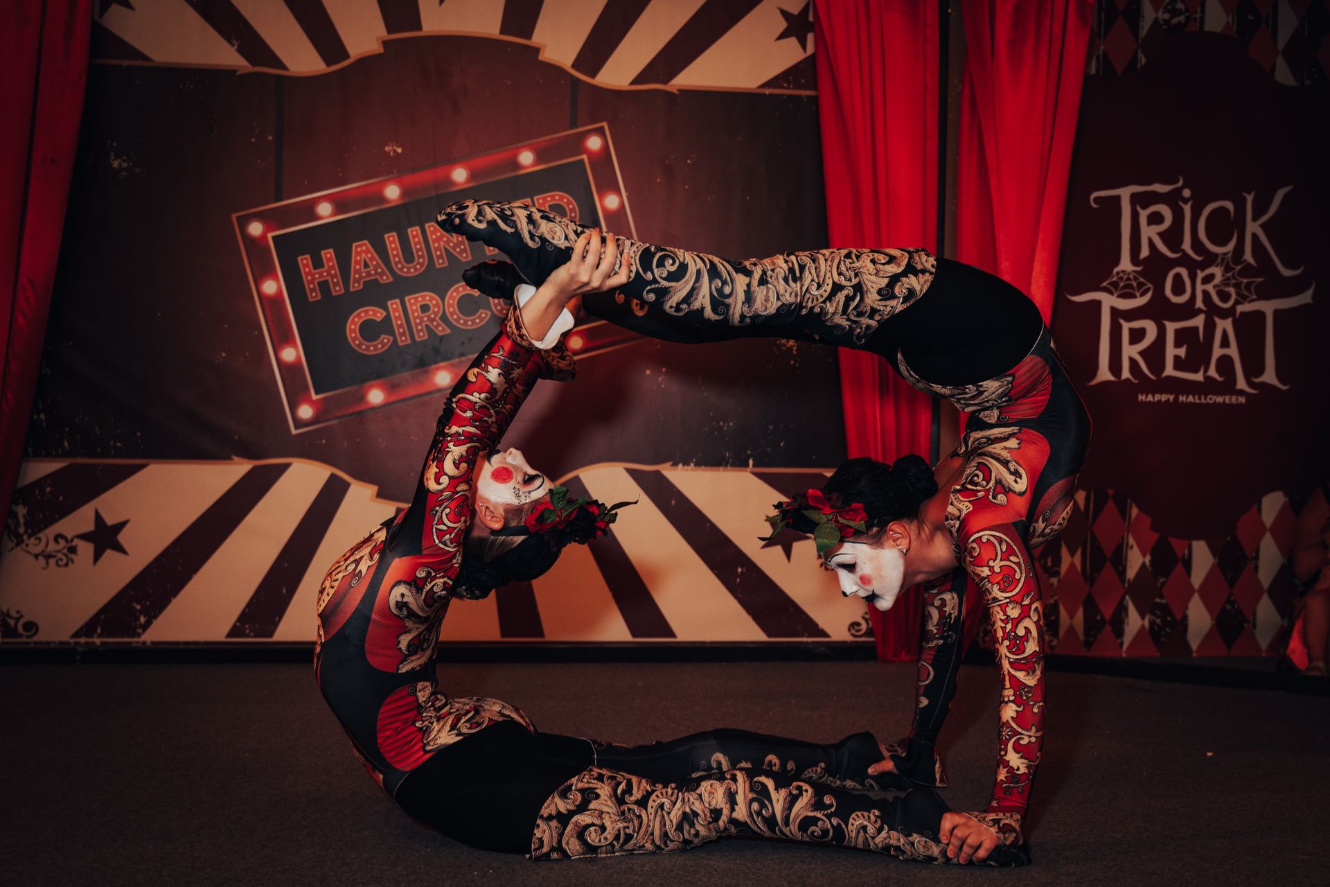 Two women perform a striking acrobatic act with intricate face paint at a haunted circus event.