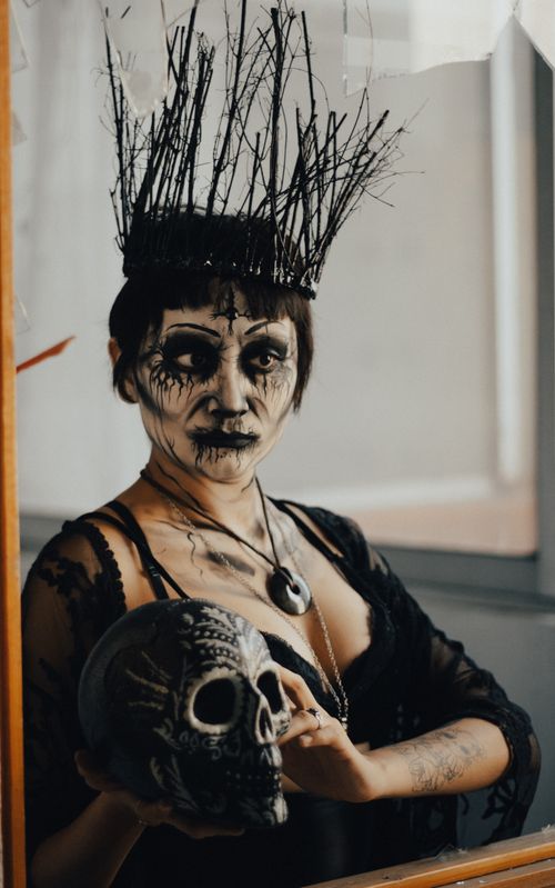 Free A woman with dramatic face paint and a witch hat holds a skull in front of a mirror. Stock Photo