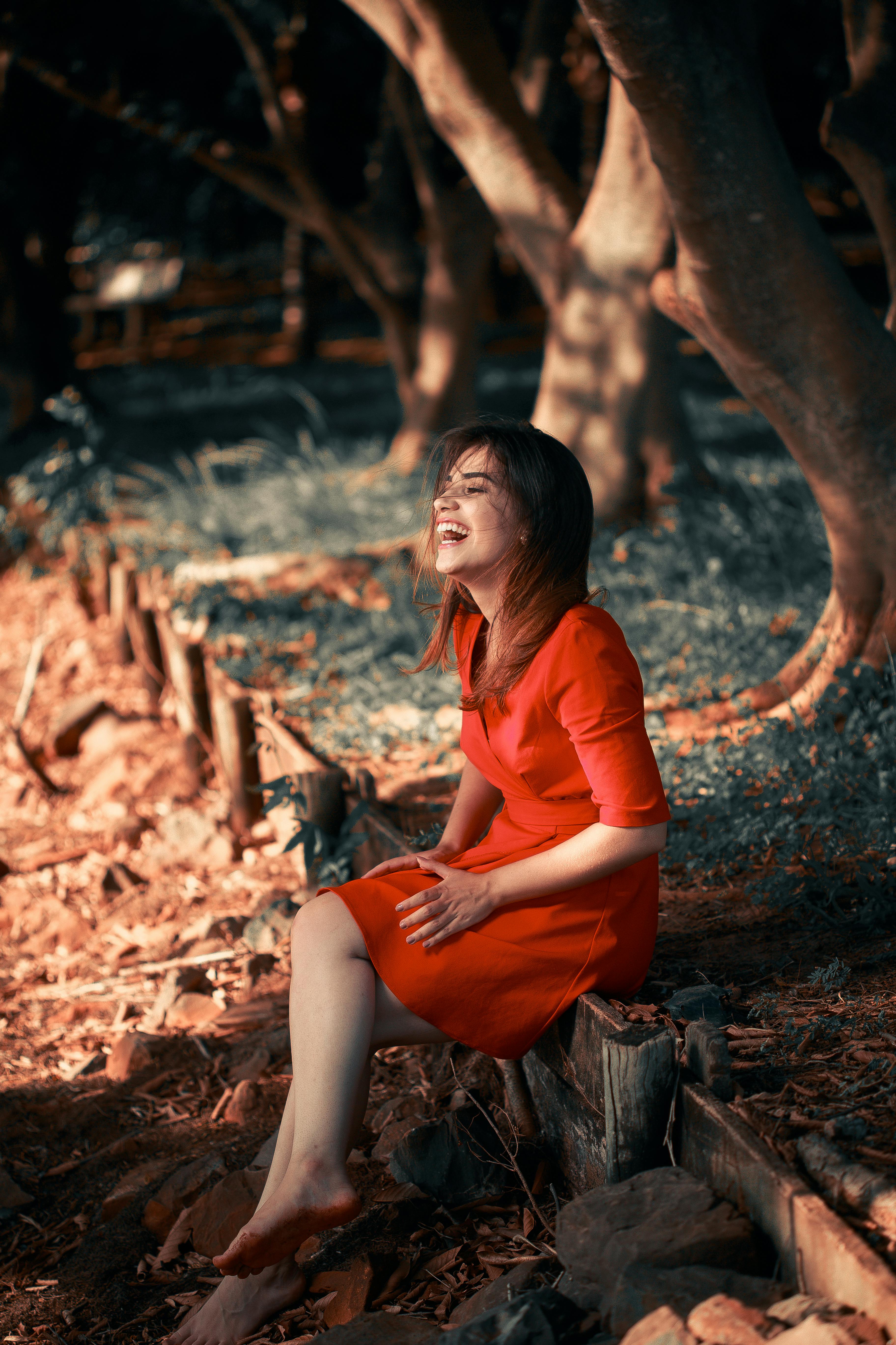 Free A woman joyfully sitting barefoot outdoors in a vibrant red dress. Stock Photo
