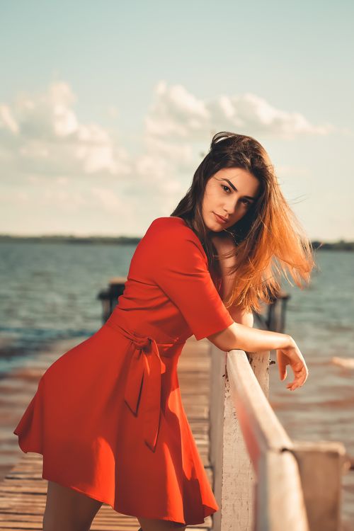 Free Beautiful woman in red dress posing gracefully on a lakeside jetty, with serene water in the background. Stock Photo
