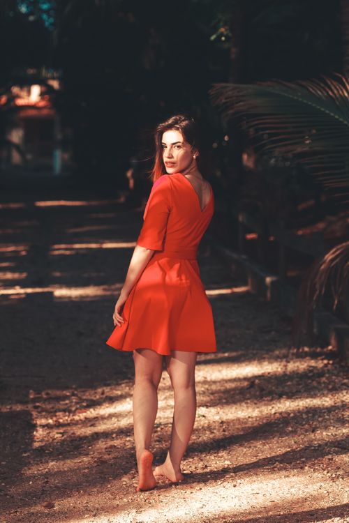 Free A woman in an orange dress walks barefoot outdoors, glancing back under dappled sunlight. Stock Photo