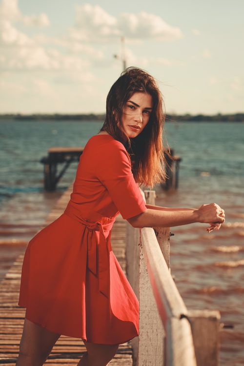 Free Fashionable woman in a vibrant red dress posing by the sea on a wooden pier. Stock Photo