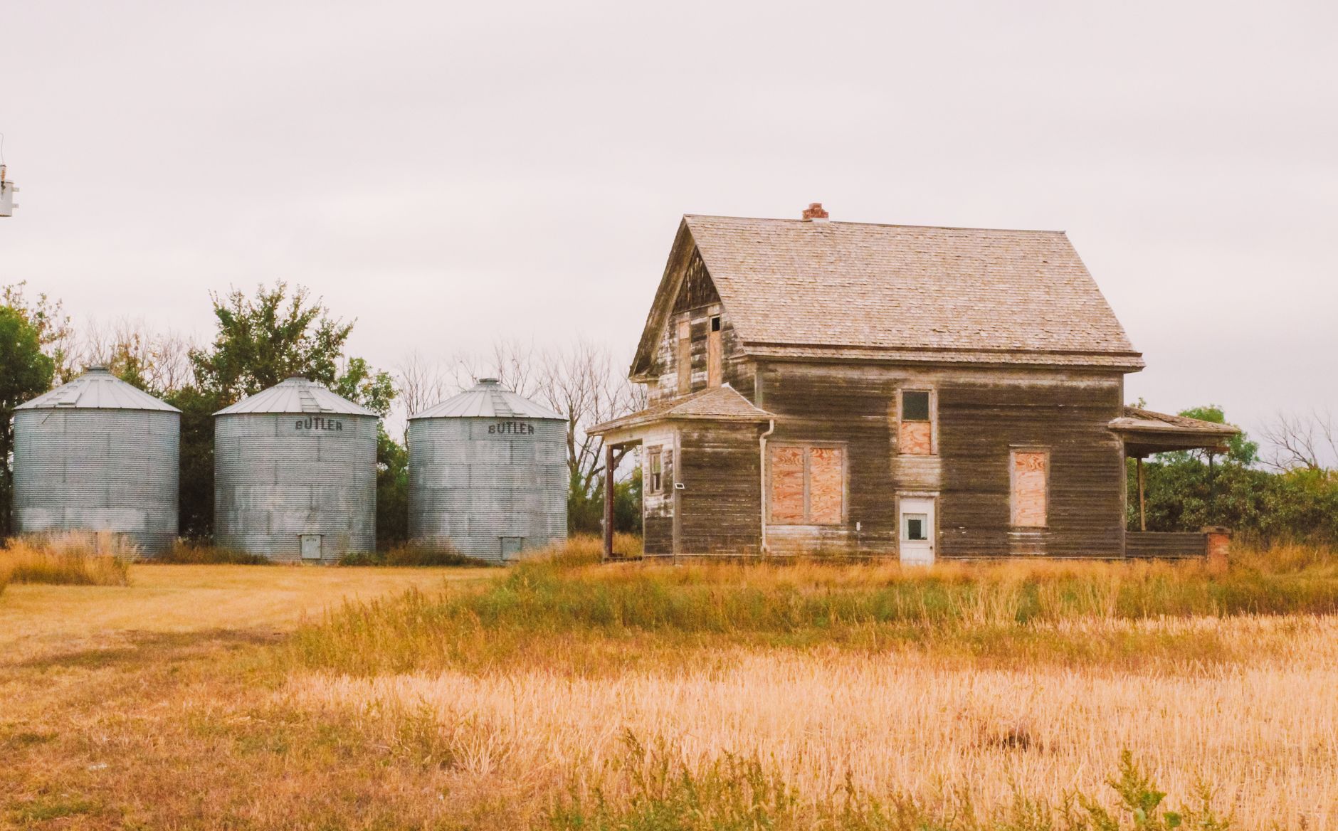 An abandoned farmhouse next to grain silos in a rural countryside setting.