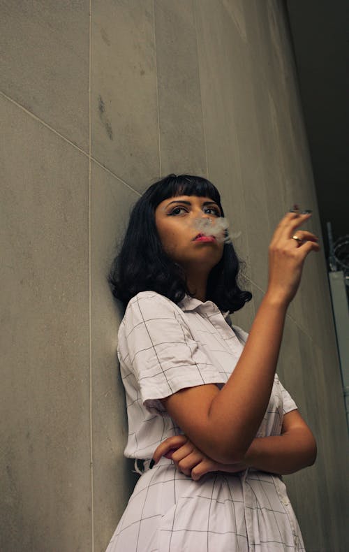 A Woman Wearing White Dress Leaning on a Gray Wall While Smoking