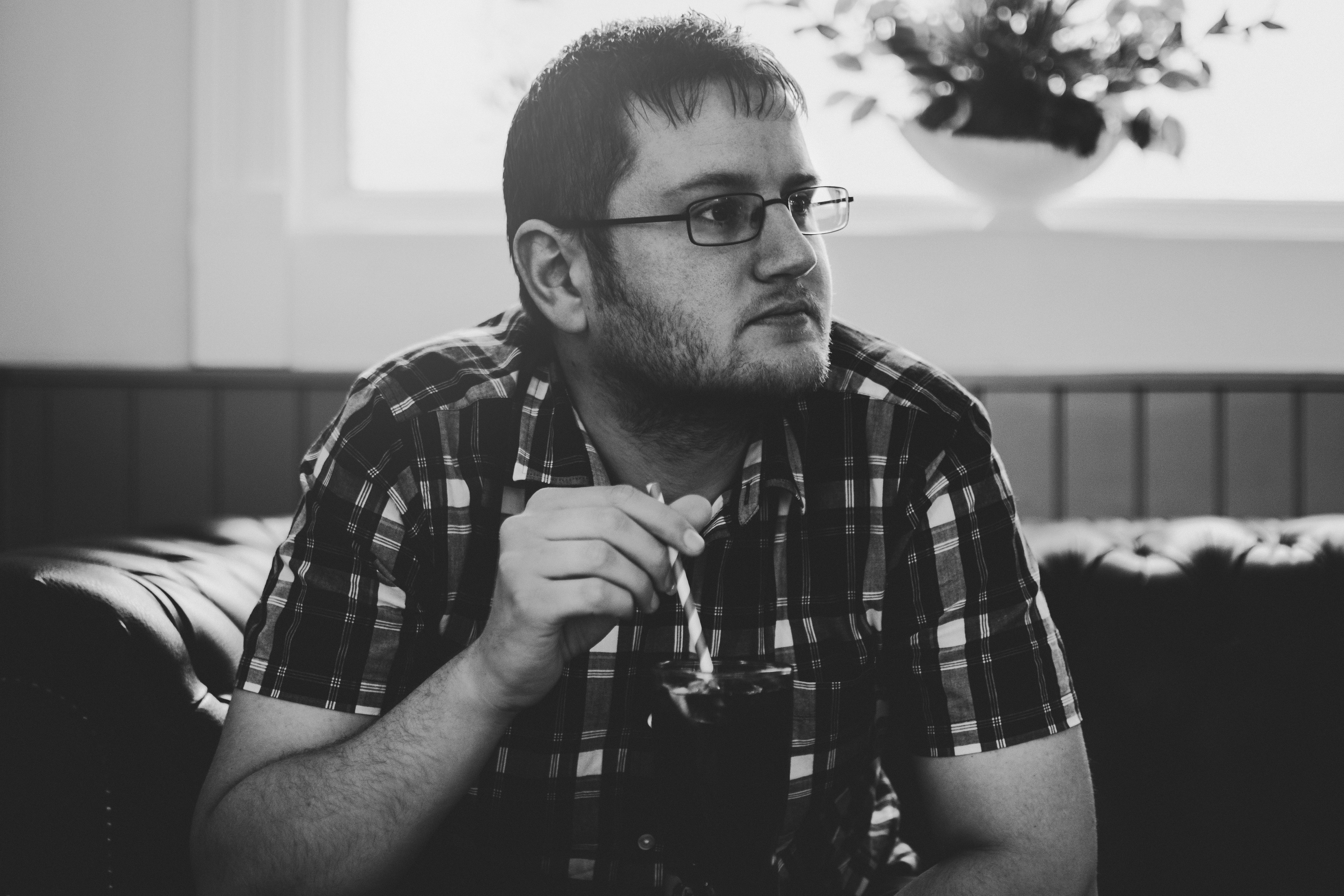 A thoughtful man in glasses enjoys a drink through a straw indoors. Black and white photo.