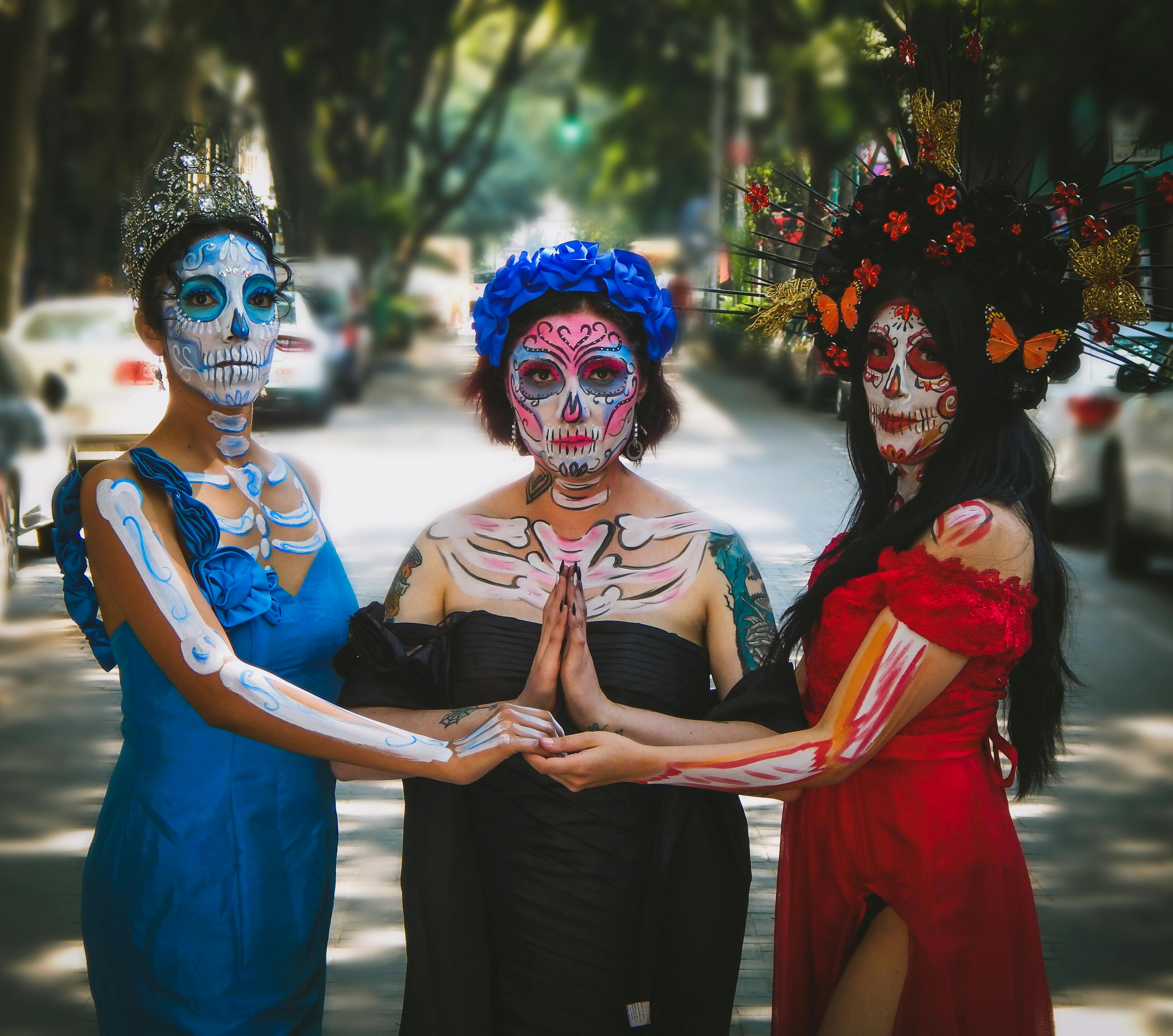 Three women celebrate Dia de los Muertos with intricate face paint and colorful dresses on a city street.