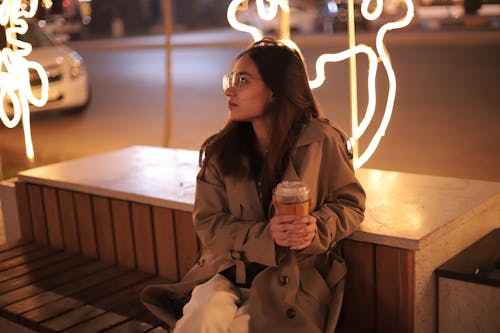 A Woman in Brown Coat Sitting on the Bench while Holding a Drink