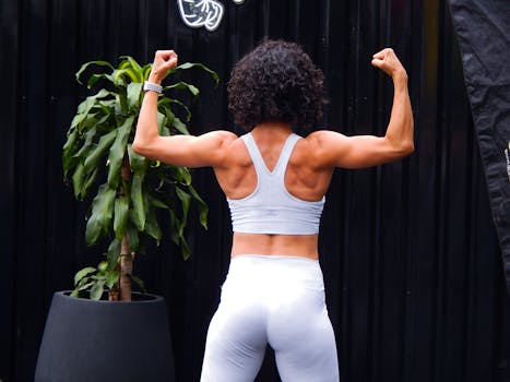 A woman in activewear flexes her muscles confidently outdoors next to a plant.