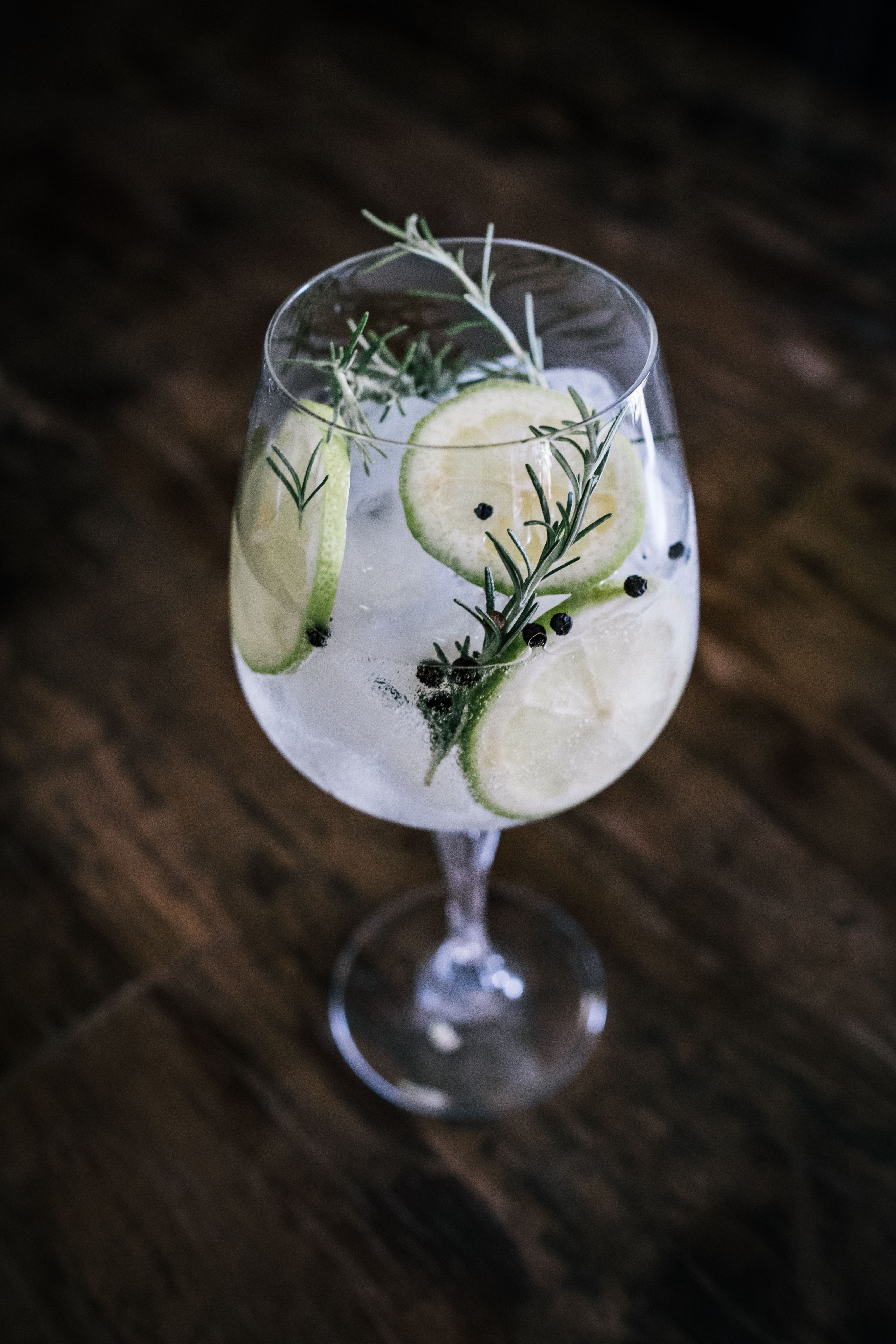 A close-up of a gin and tonic garnished with lime slices, rosemary and juniper berries, in a glass.