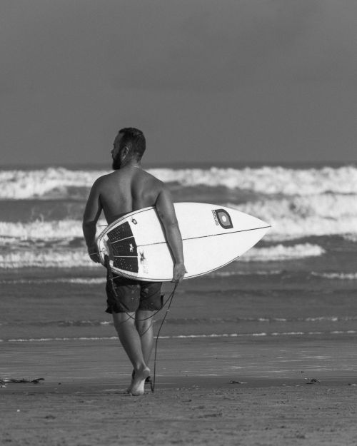 Free A man walks on a beach holding a surfboard, captured in black and white, embodying a serene coastal vibe. Stock Photo
