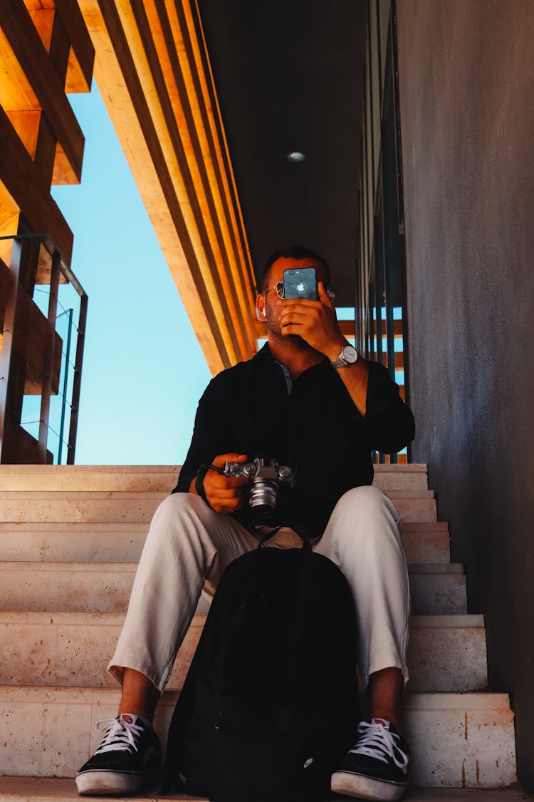 A Man In Black Long Sleeves Sitting On The Stairs While Holding His Phone And Camera