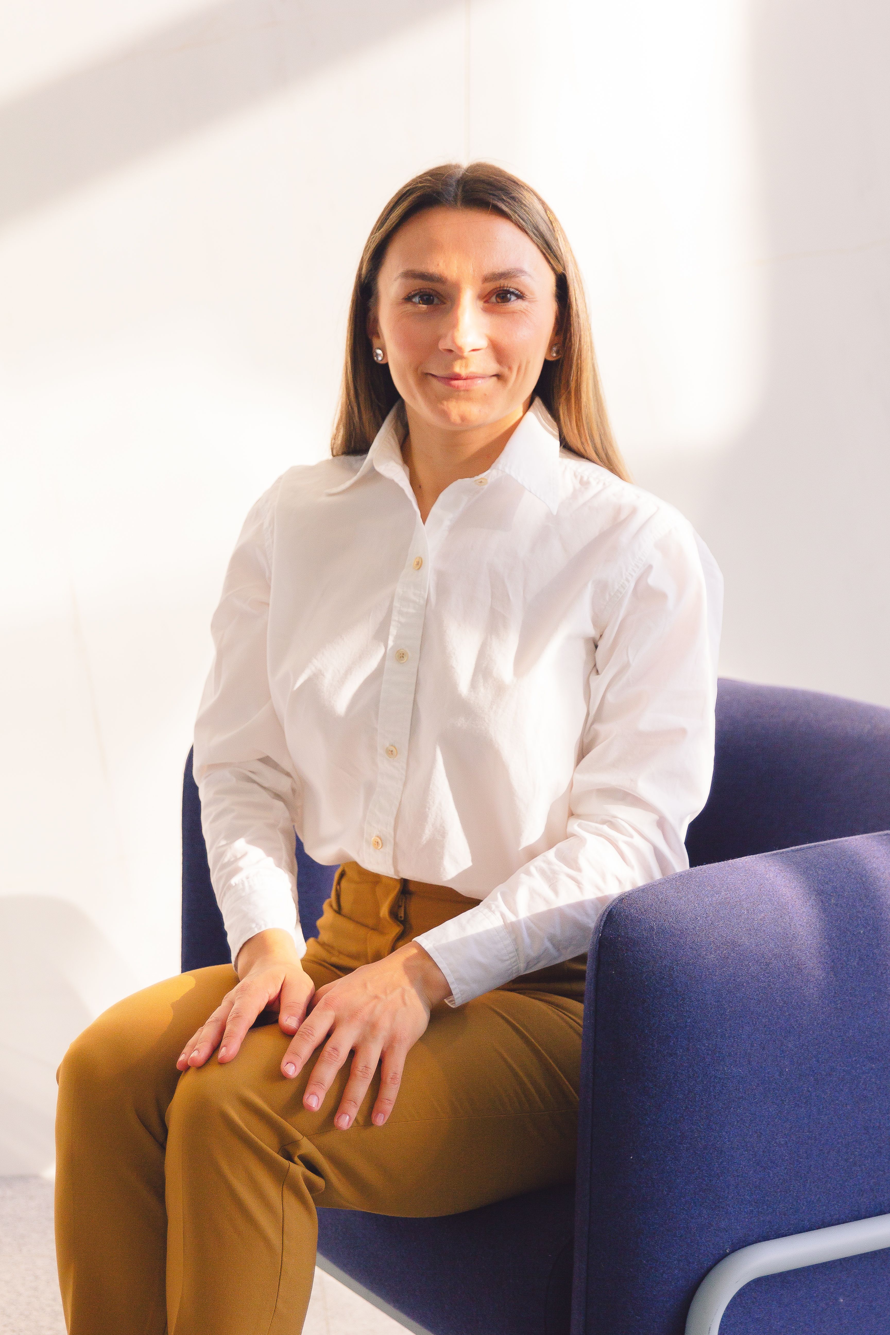 Elegant woman in white shirt sitting on a blue chair in bright light.