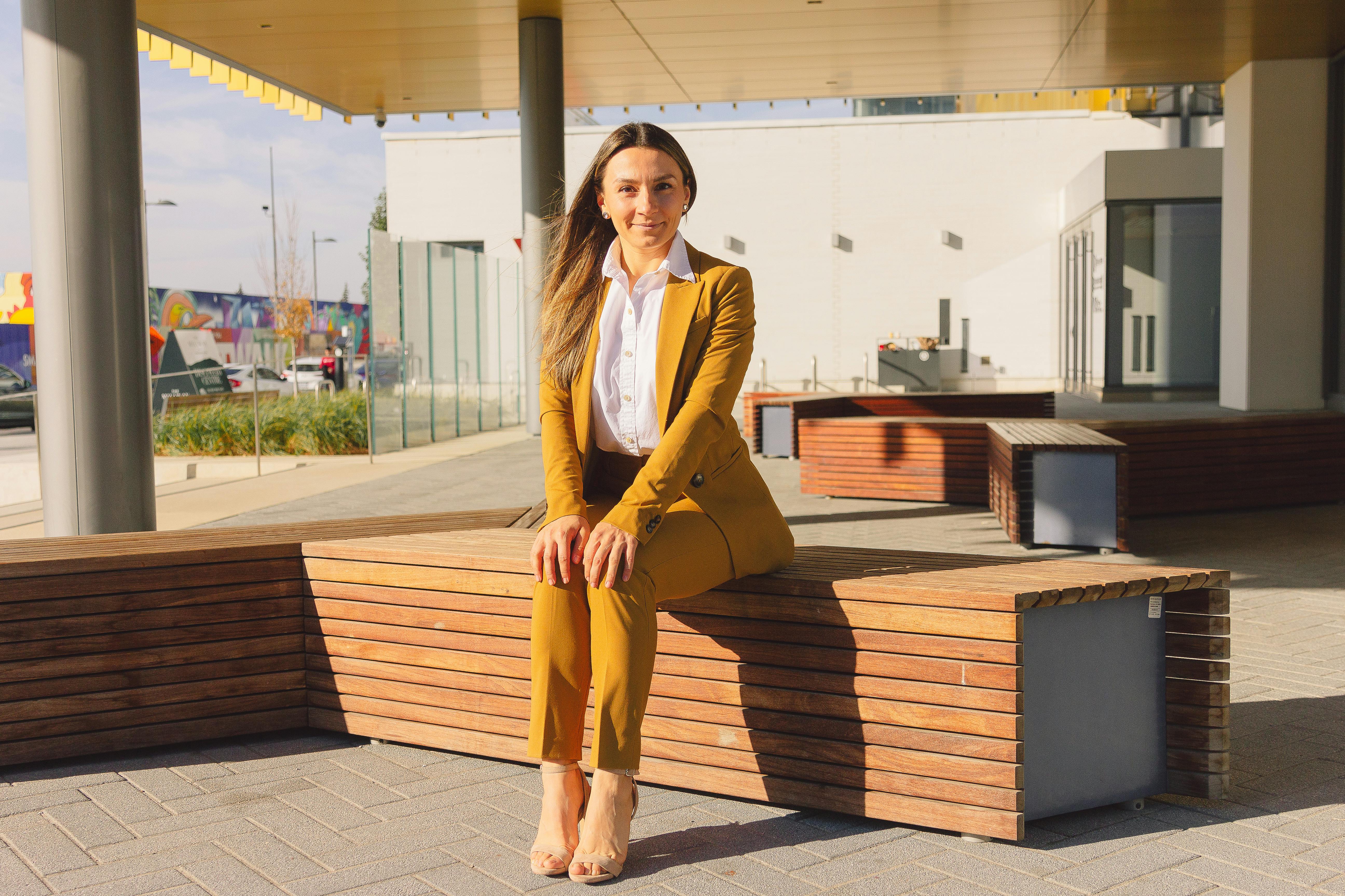 Woman in a yellow blazer and pants sitting on a wooden bench, smiling confidently.