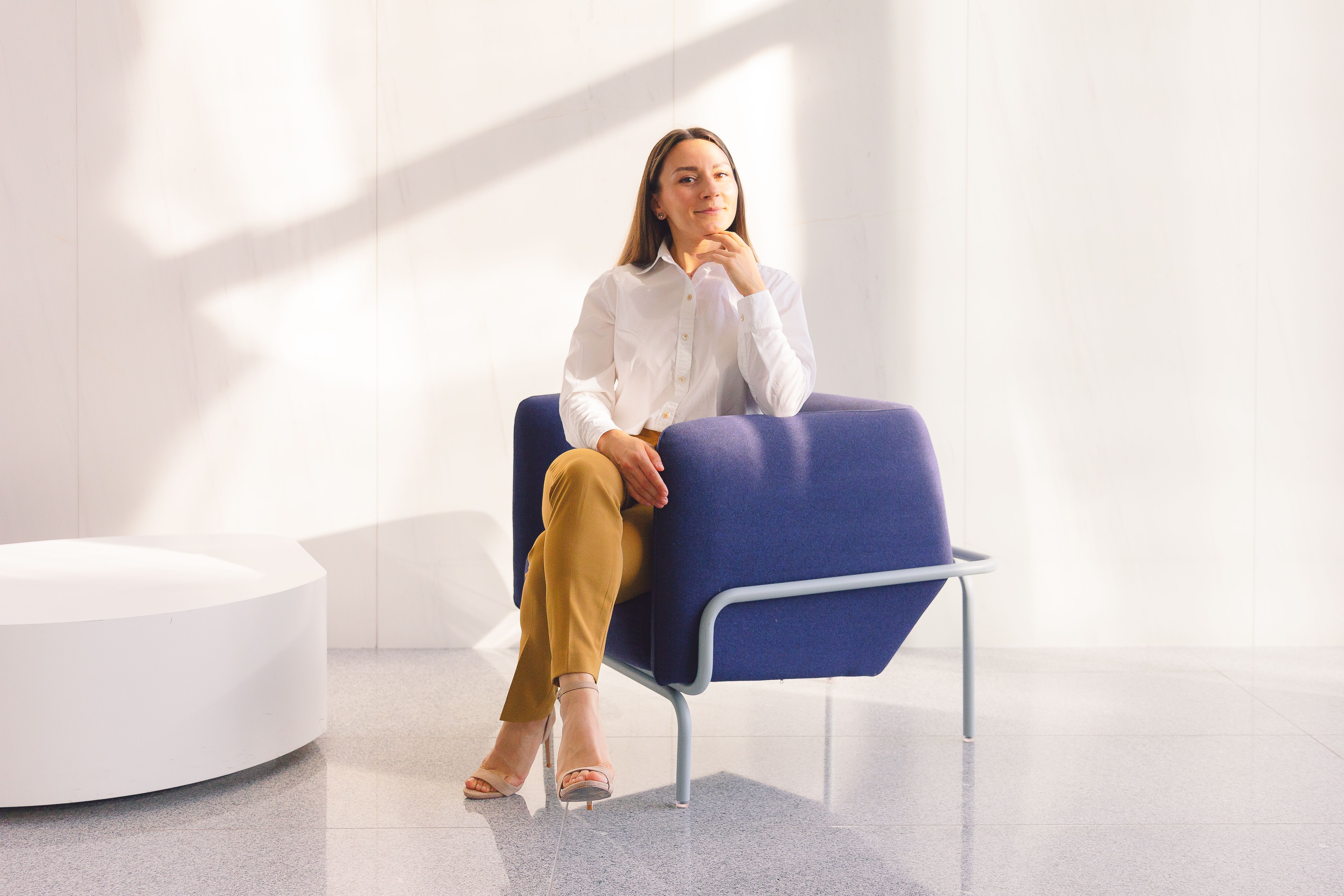 Confident woman in a white shirt and yellow pants sitting on a stylish blue chair in a bright modern space. | BocaVibe