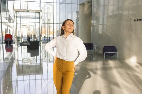 Free Young woman in white blouse and brown pants confidently posing in a bright modern office lobby. Stock Photo